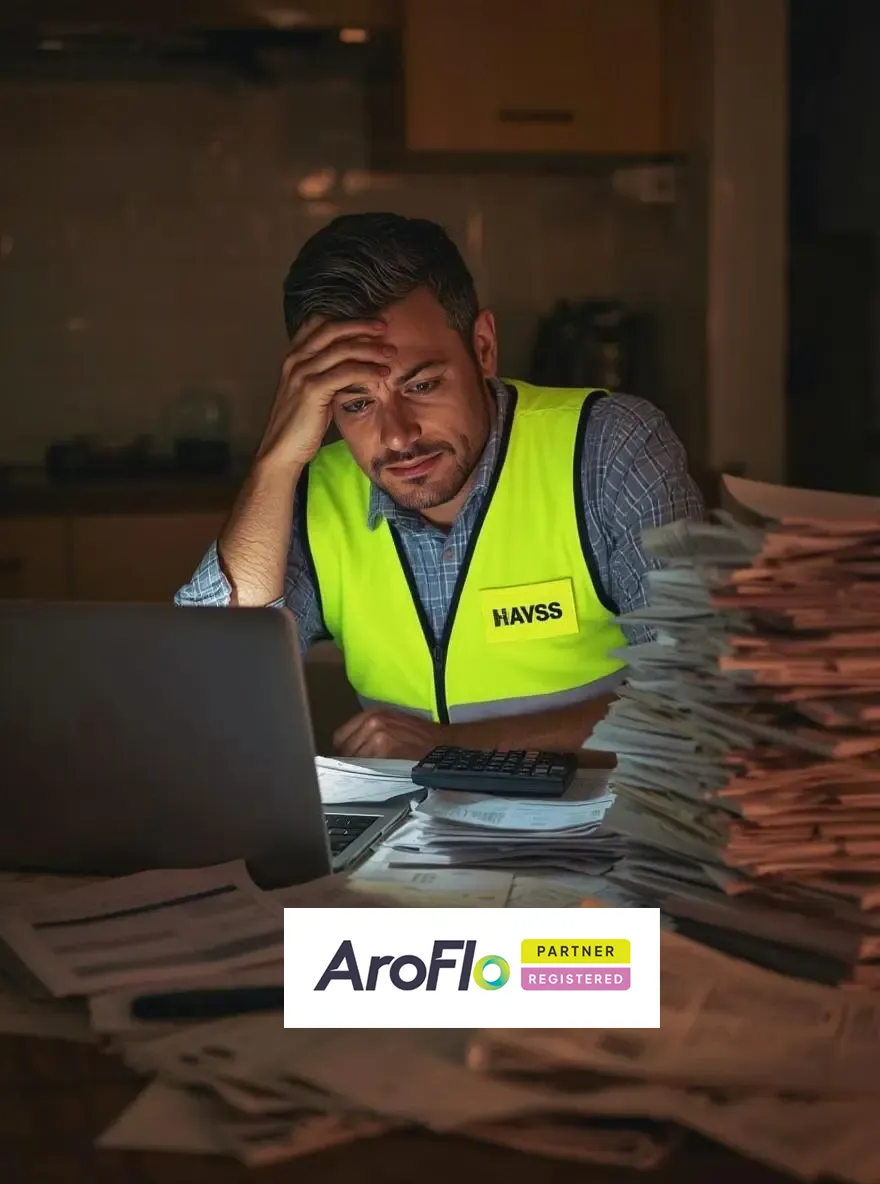 Stressed man in high-vis vest working on a laptop surrounded by piles of paperwork.